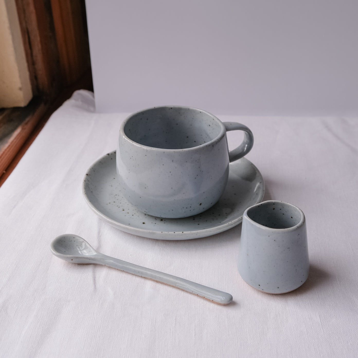 A blue pepper speckled tea and coffee mug set with a matching saucer, teaspoon and milk pourer by Palinopsia Ceramics on a white linen tablecloth in the boutique ceramics store in Newcastle Australia