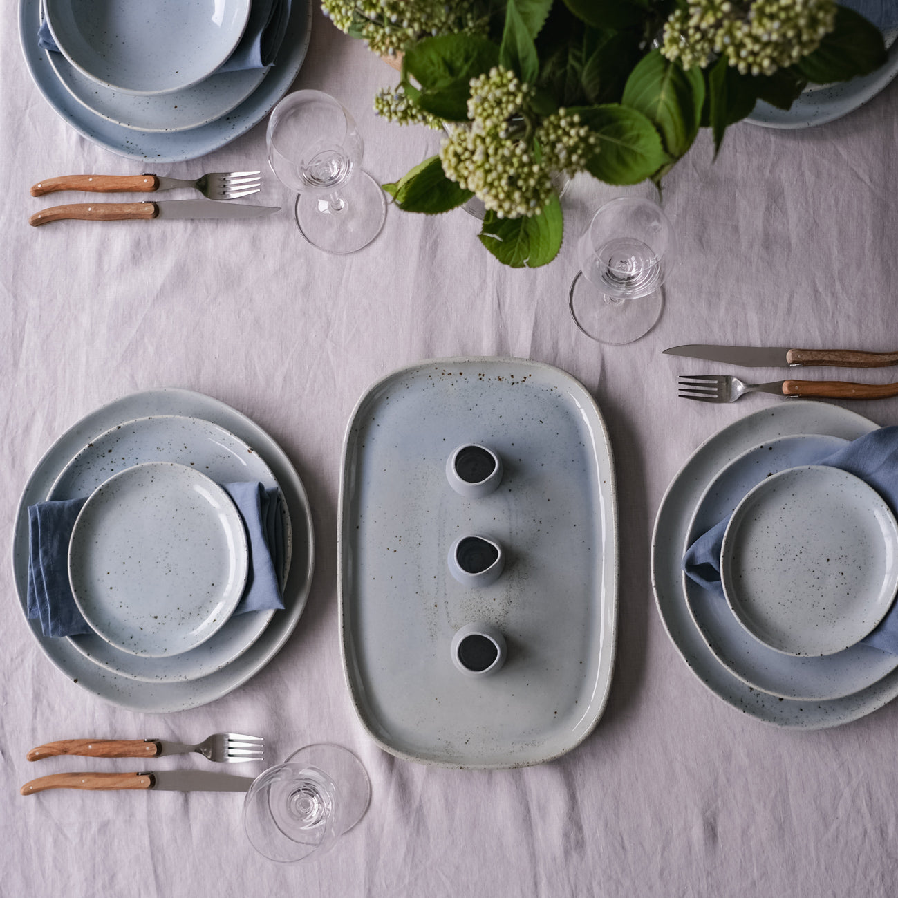 Handmade little gravy jugs in blue speckle on a dressed table with a grey linen tablecloth and blue dinnerware.